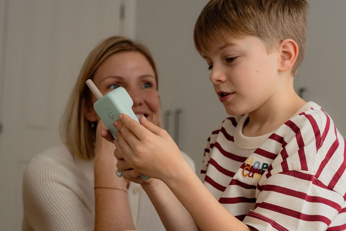 A boy holding a mint green Waffle Home handset to his ear while his mum sits beside him.