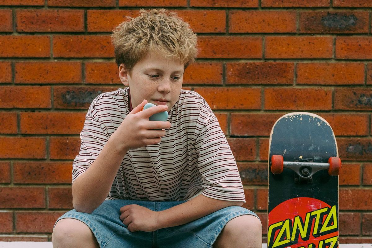 A boy sitting against a brick wall with his skateboard, speaking into a mint green Waffle Go phone.